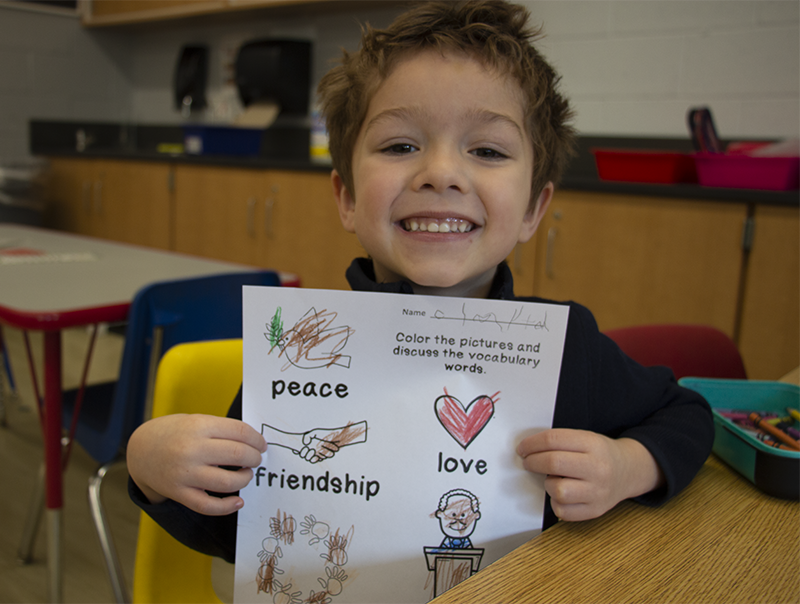 A pre-K student smiles as he holds up a paper he colored about Martin Luther King.