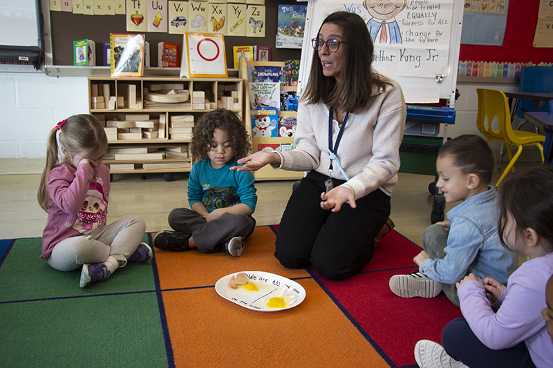 A woman with long dark hair and glasses kneels on a colorful rug with young pre-K students. In front of her is a plate with two eggs broken onto it.