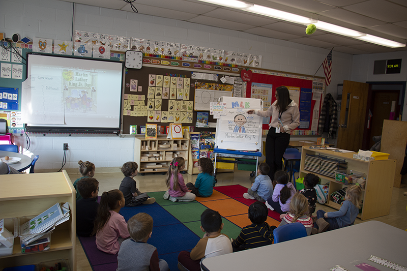 A woman with long dark hair and glasses stands in front of a group of pre-K students, pointing to a board that has a picture of Martin Luther King Jr on it.