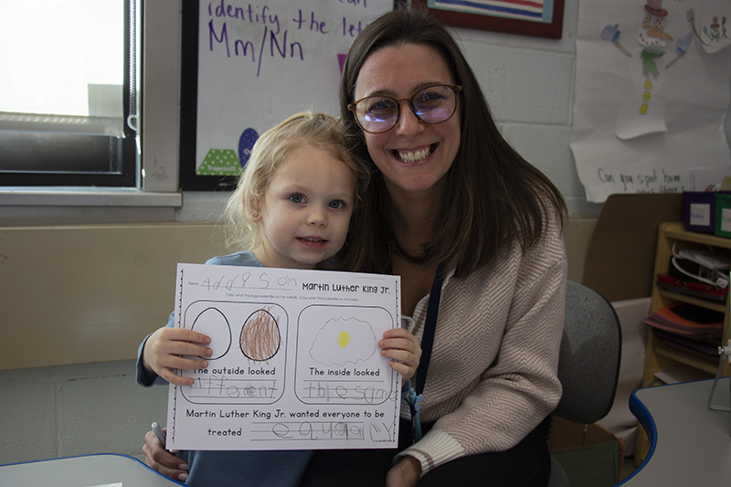 A pre-K girl smiles and holds a piece of paper she has colored and written on. She is with a woman with long dark hair and glasses who is also smiling.