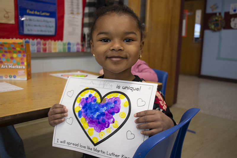 A young pre-K student smiles and holds up a heart that she decorated with blue purple and yellow markers.