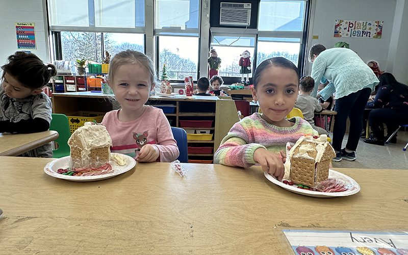 Two small girls smile with gingerbread houses they just made in front of them.