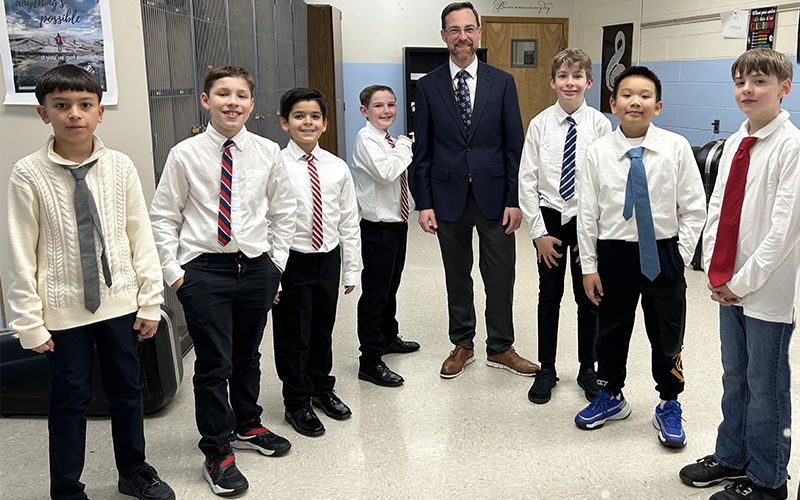 A man with dark hair, wearing glasses and a suit and tie smiles as he stands with seven older elementary students, all wearing shirts and ties.