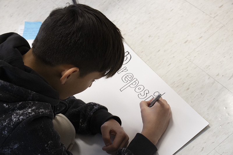 A middle school students draws on a poster board, writing Prepositions in large letters.