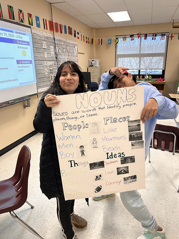 Two middle school students hold a poster that lists and shows pictures of nouns. 