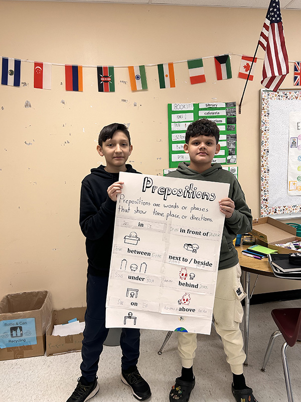 Two middle school students hold a large poster with the heading prepositions and lists of words on it.