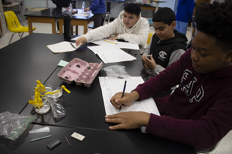 A group of three sixth-grade students, all wearing goggles, writing on paper after doing an experiment. There is a pink egg carton on the table with eight minerals  in it.