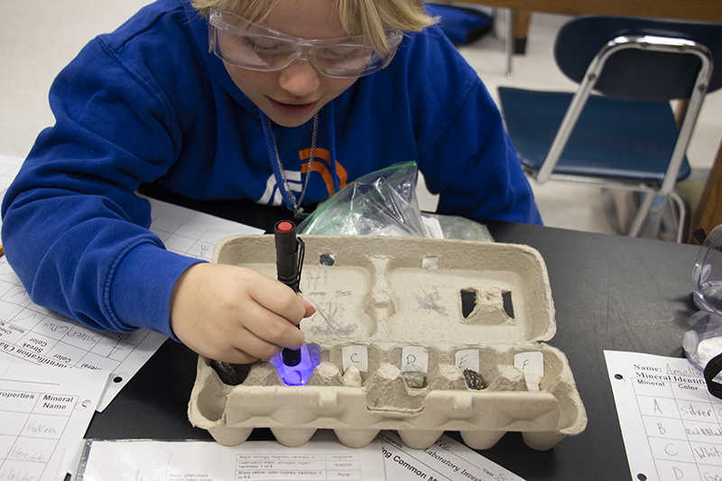 A boy with blonde hair and wearing safety goggles uses a fluorescent light to see properties on small minerals.