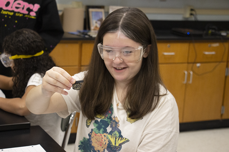 A girl with long dark hair, wearing safety goggles, smiles as the  mineral she is testing sticks to a magnet.