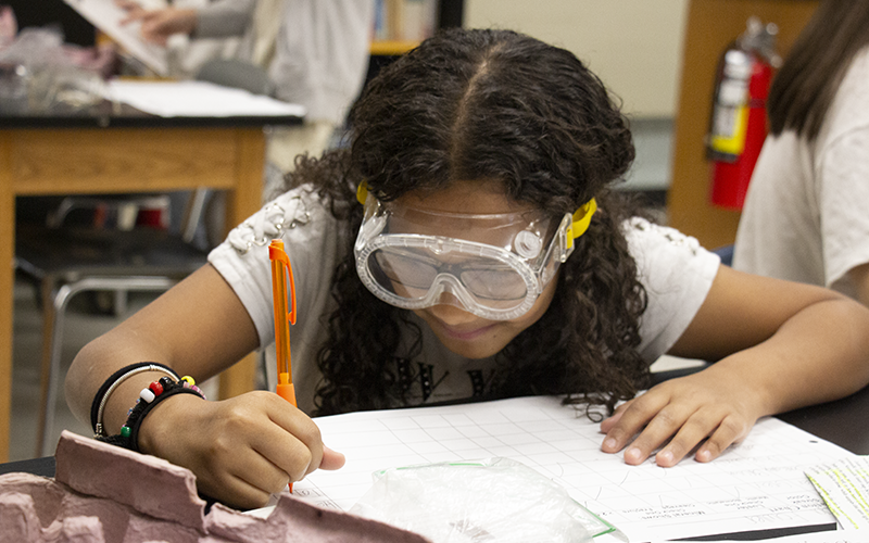 A girl with long dark hair, wearing safety goggles, writes her findings on a piece of paper.