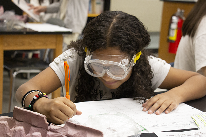 A girl with long dark hair, wearing safety goggles, writes her findings on a piece of paper.