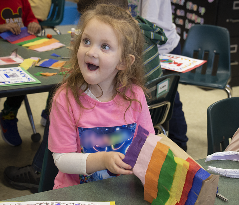 A little girl with long blonde hair gives a very surprised happy look to someone as she makes a colorful pinata.