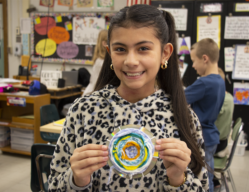 A girl with long dark hair  smiles and holds up a CD on which she has weaved a pattern with yarn.