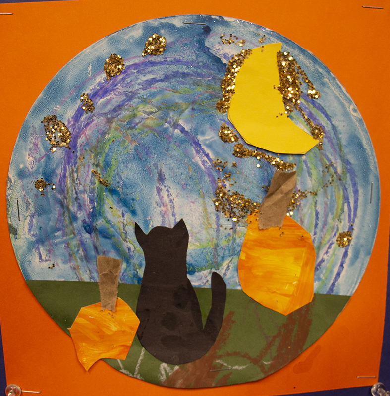 A round drawing using cut out moon, black cat and pumpkins. The sky is colored blue.