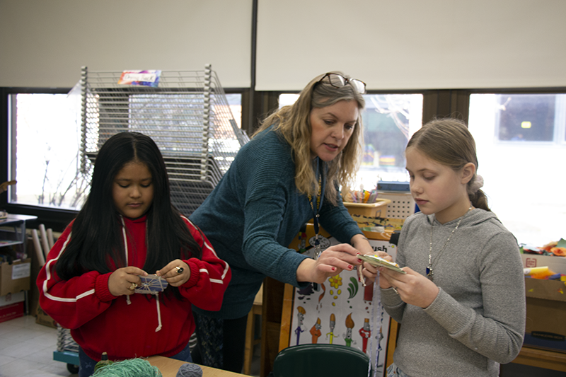 A woman with long blonde hair leans in to help a fourth-grade student with a weaving project. A girl stands next to them working on hers.