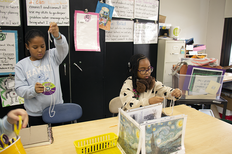 Two fourth-grade girls weave a project in their classroom, using yarn and a CD.