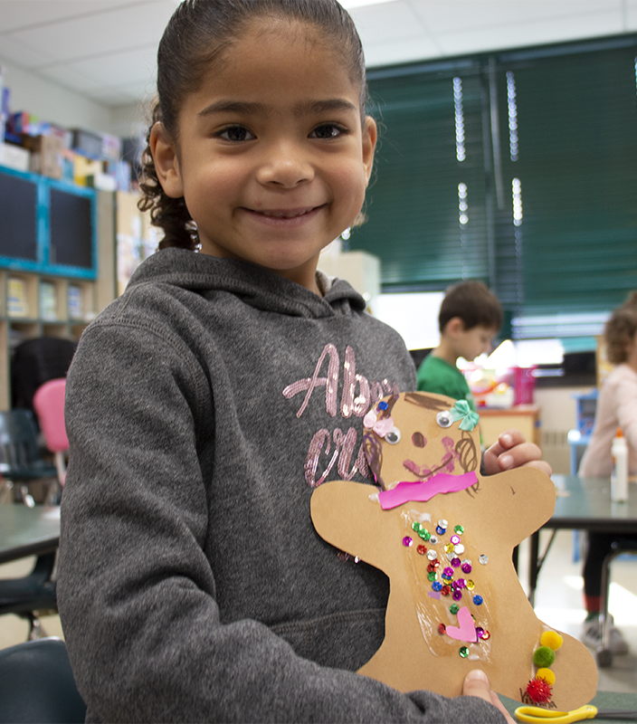 A girl with dark hair pulled back smiles. She is holding a gingerbread woman she made from paper and decorated with colorful beads and paper.