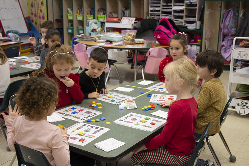 A group of six kindergarten students sit at a table  where they are playing bingo with brightly colored markers.