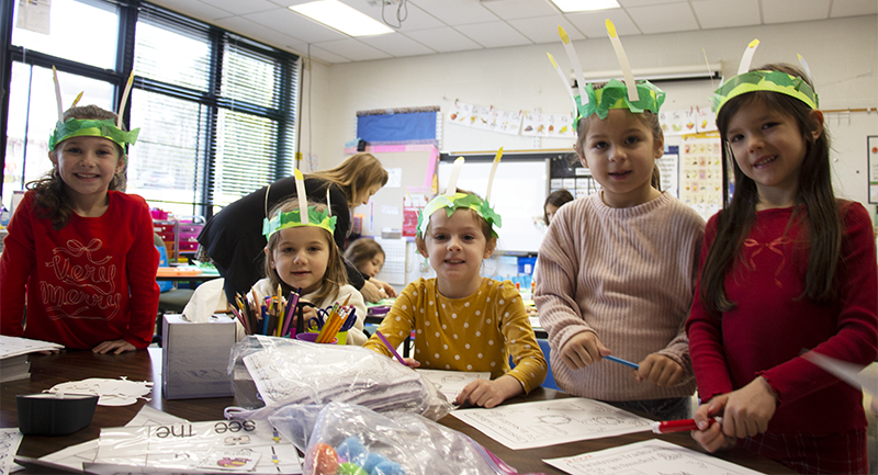 Five little kindergarten girls smile as they wear green crowns with paper candles in them, which they made.