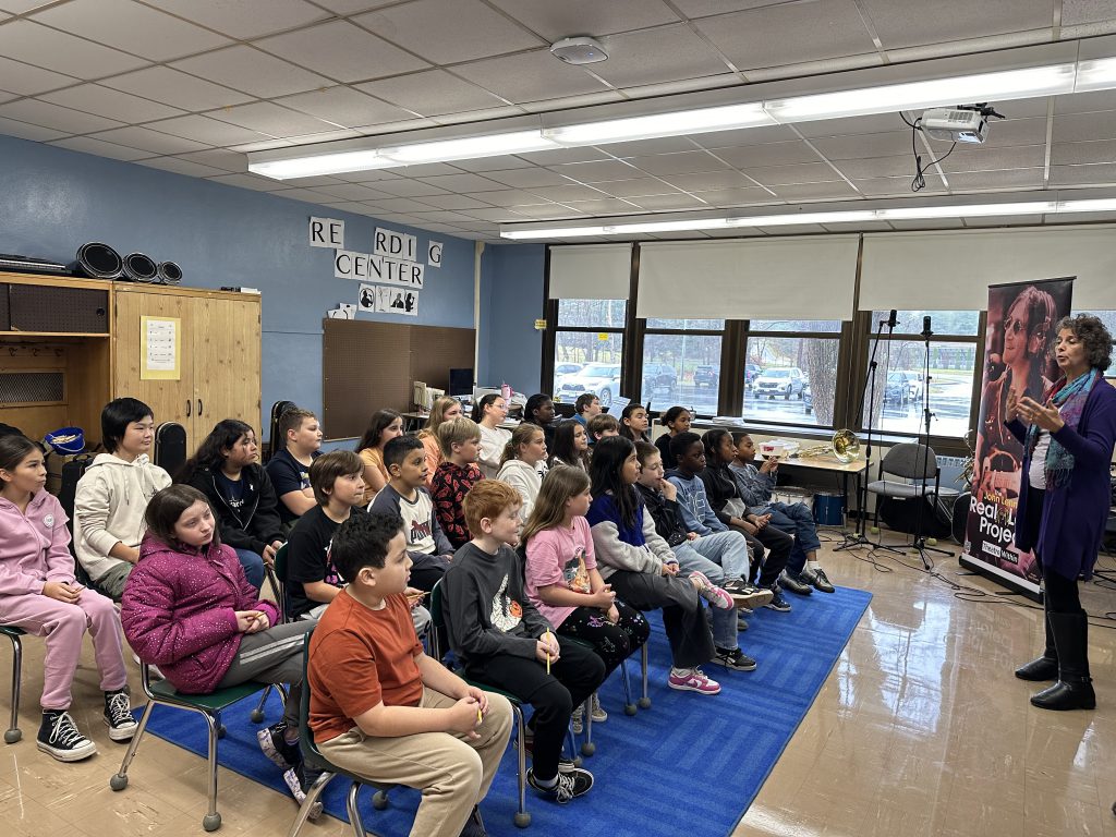 A group of about 25 fifth-grade students sit and listen as a man in front of a classroom directs them.