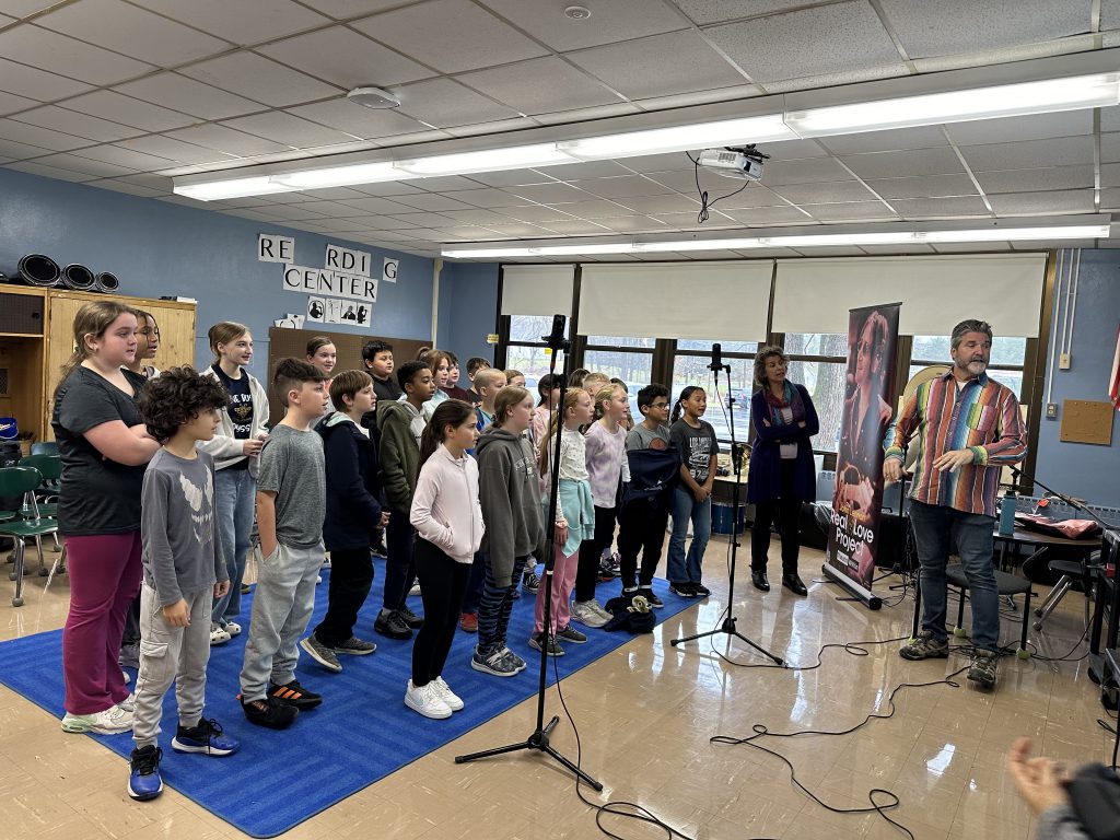 A group of fifth-grade students all stand around a microphone as they record a song.