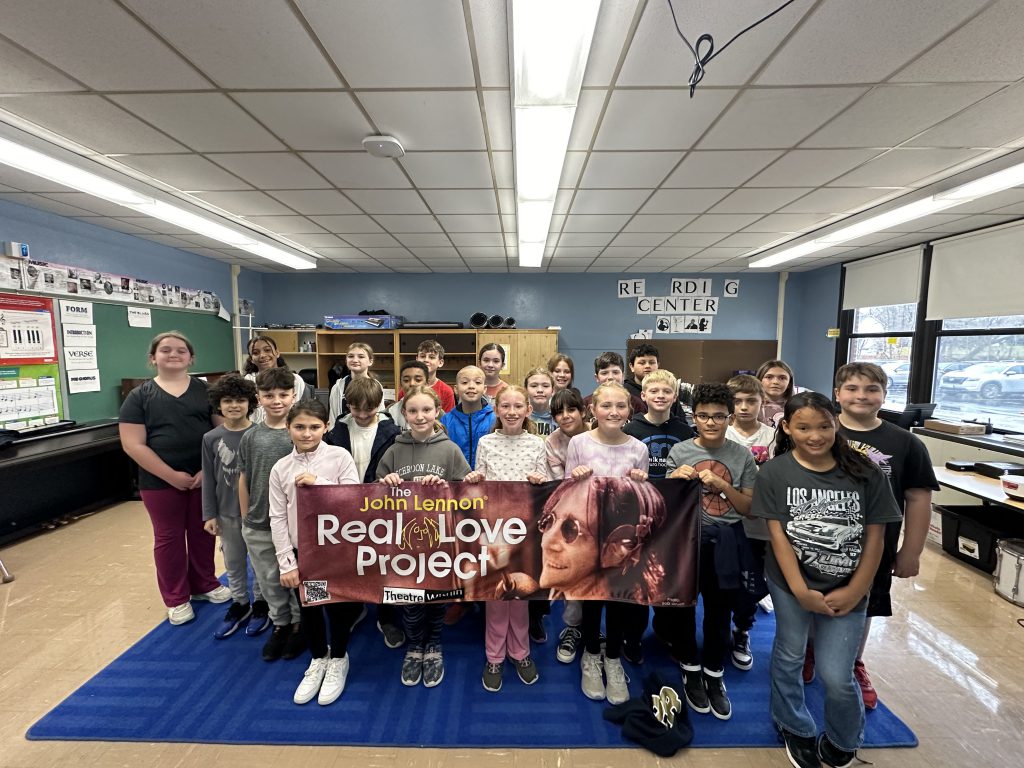 A large group of fifth-grade students  stand and smile. The few in front hold a banner that says John Lennon Real Love Project.