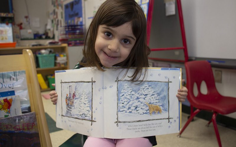 A little girl with long dark hair holds a book open and smiles. The book has pictures of winter in it.