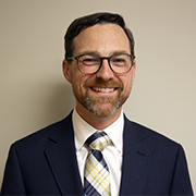 A man in a navy blue suit, white shirt and blue and gold tie, wearing glasses smiles. He has short dark hair and a close cut beard and mustache.