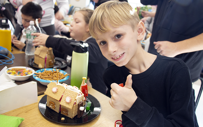 A boy with blonde hair gives a thumbs up. He is sitting next to his gingerbread house he made.