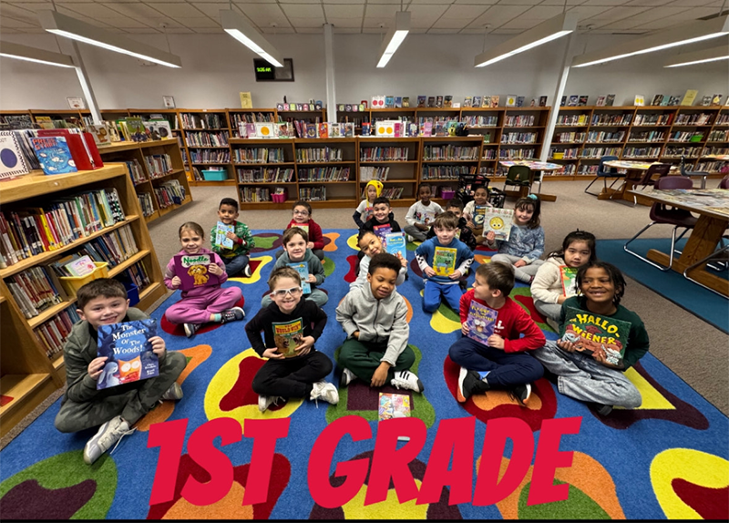 A group of 17 first grade students sit on a colorful carpet, each holding a book. They are in a library.