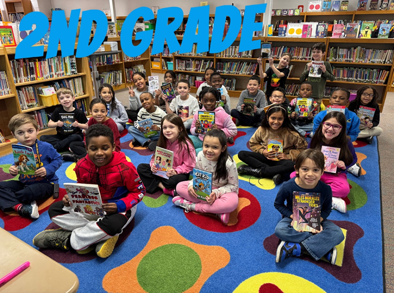 A group of 24 second grade students sit on a colorful carpet, each holding a book. They are in a library.