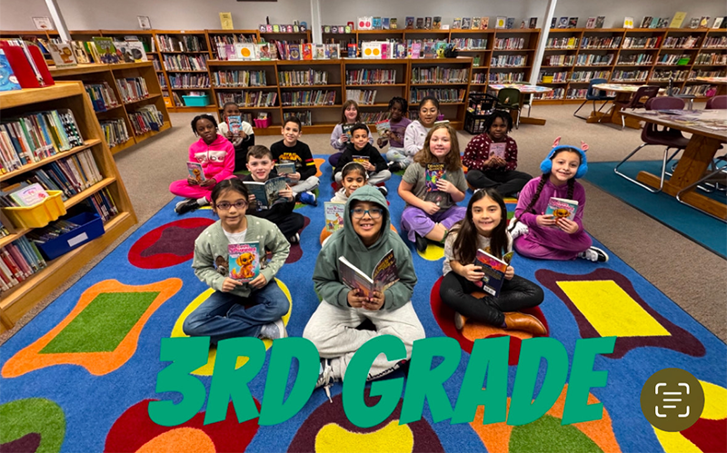 A group of 15 third grade students sit on a colorful carpet, each holding a book. They are in a library.