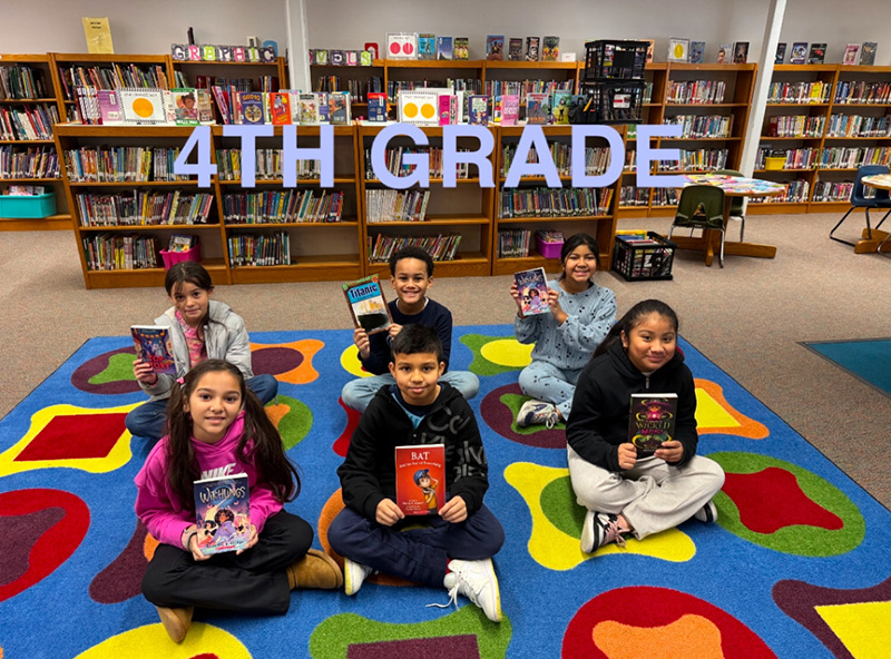 A group of six fourth grade students sit on a colorful carpet, each holding a book. They are in a library.