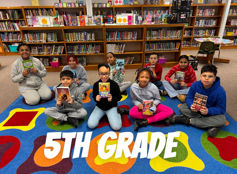 A group of 9 fifth grade students sit on a colorful carpet, each holding a book. They are in a library.