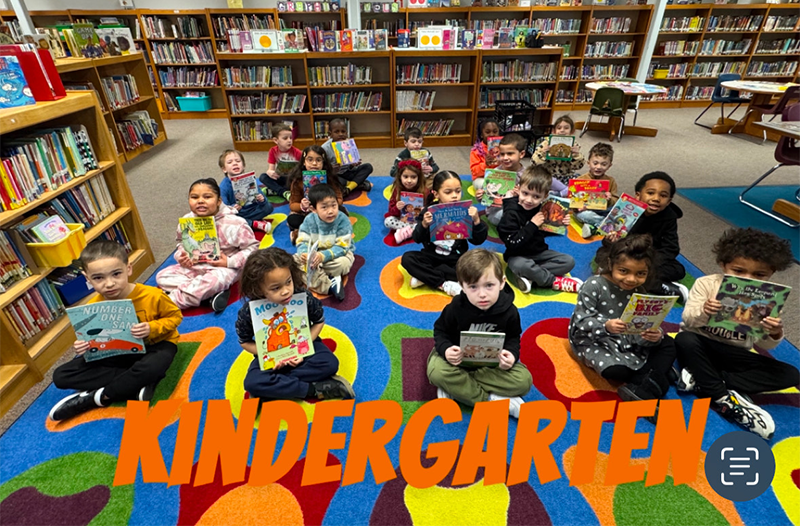 A group of 20 kindergarten students sit on a colorful carpet, each holding a book. They are in a library.