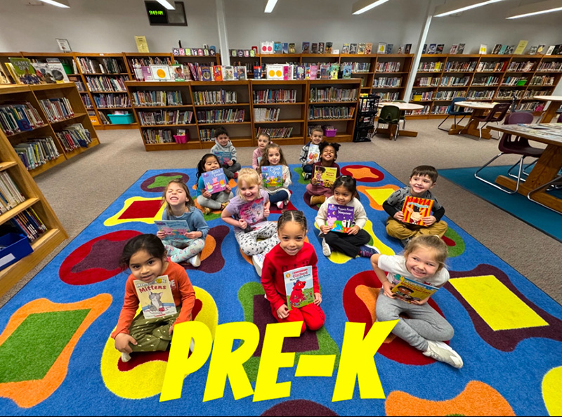 A group of 13 pre-K students sit on a colorful carpet, each holding a book. They are in a library.