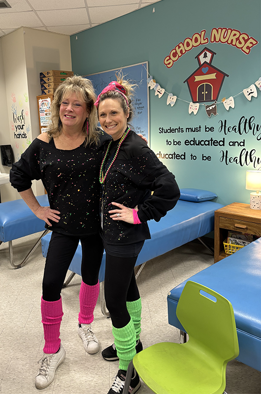 Two adult women with their hair up big, wear all black with splatters of neon colors  on them. They are wearing slouchy neon socks.