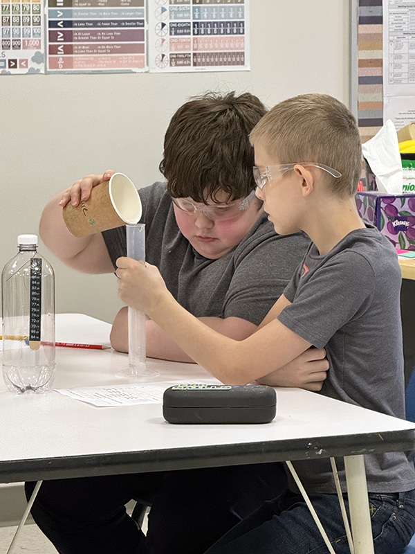 Two elementary age boys work together on a science experiment One holds a clear tube and the other pours a liquid in it.