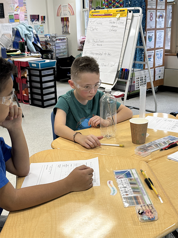 Two elementary age boys sit at a table and work on a science experiment with a clear plastic bottle.