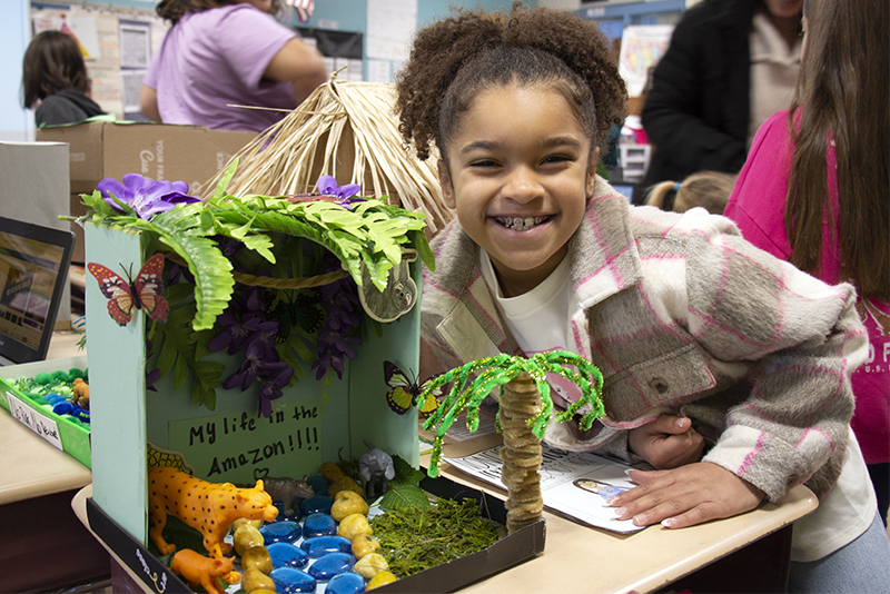 A fourth-grade girl leans over a desk that has a diorama she made, with palm trees and animals. She is smiling broadly.