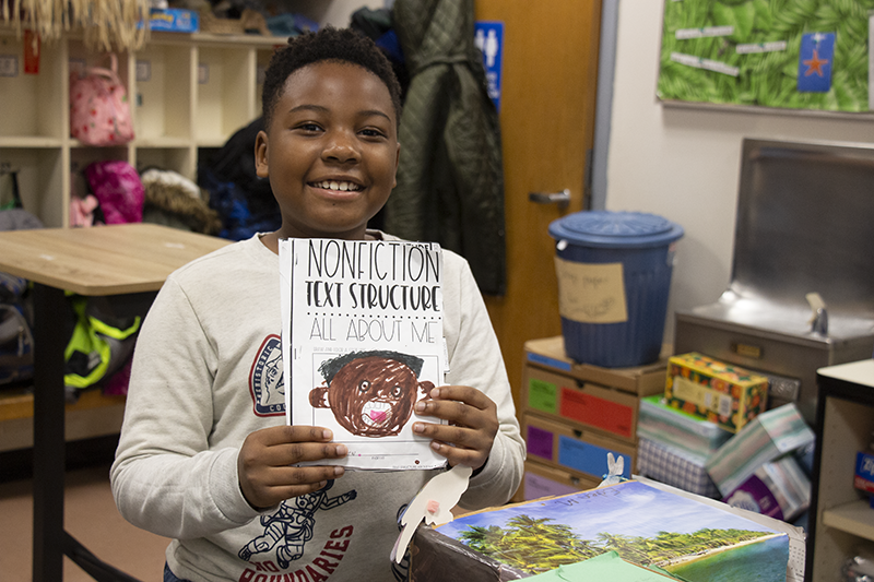 A fourth-grade boy smiles as he holds up a booklet he wrote about himself. he drew a picture of himself. It says Nonfiction text structure All About Me