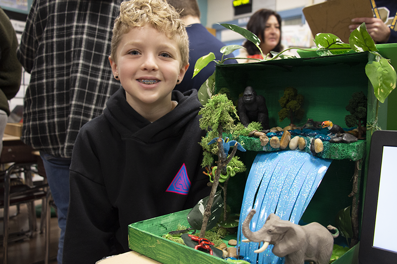 A fourth-grade boy with short curly blonde hair smiles next to the diorama he made. It has a big blue waterfall and animals.