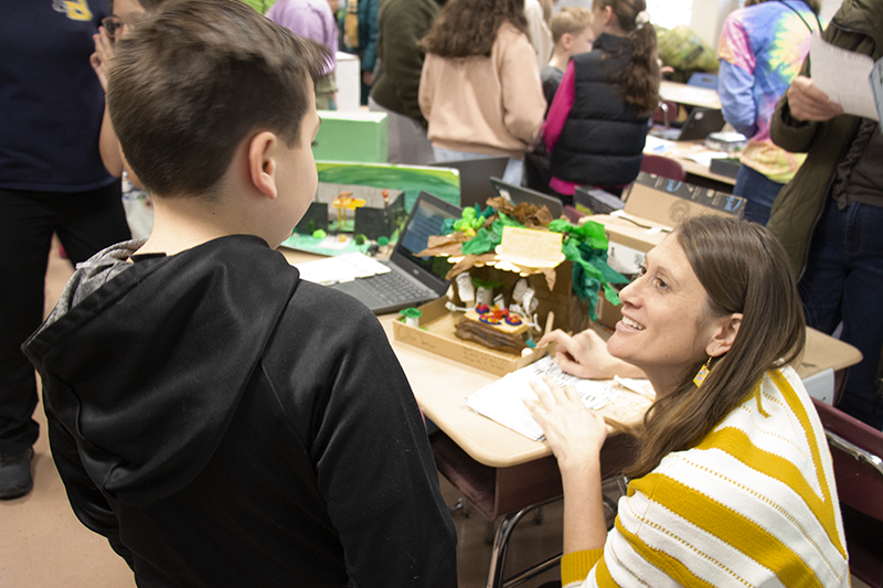 A woman with long hair squats down near a desk to talk to one of her students, a fourth-grade boy. On the desk is his diorama that he built depicting a scene in the Amazon.