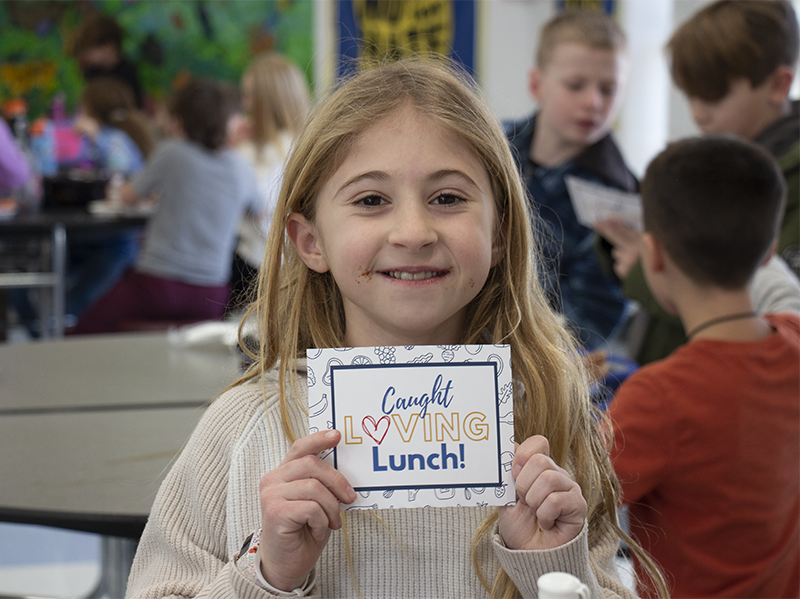 A girl with long blonde hair smiles and holds up a card that says Caught Loving Lunch!