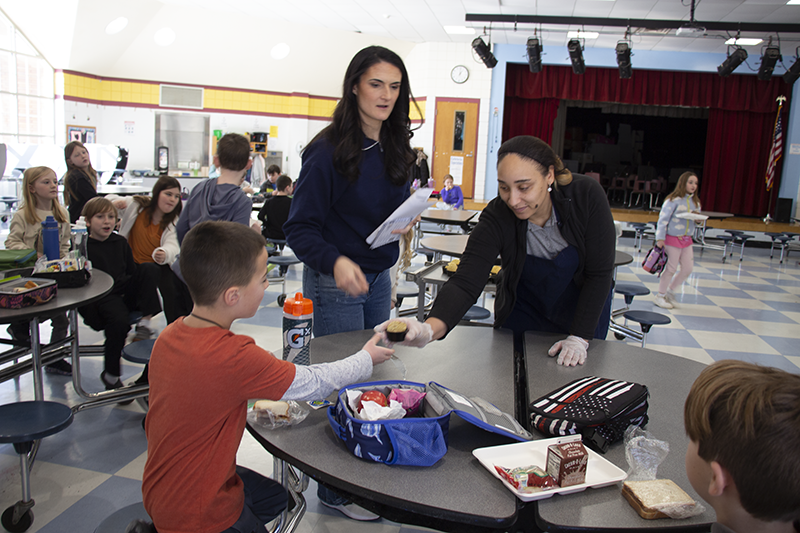 Two women with dark hair hand samples of food to an elementary kid sitting down at a table in a cafeteria.