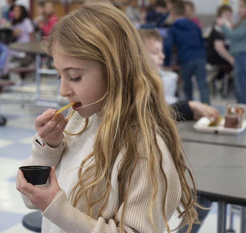 A girl with long blonde hair eats from a little sample cup.