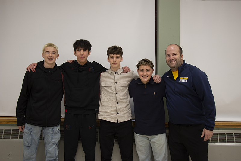 Four high school boys and a man on the right stand together, arms around each other's shoulders for a picture. They are all smiling.