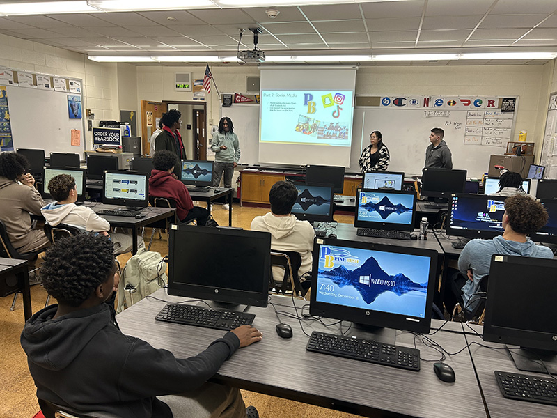 A group of high school students sit at their desks as other students are at the front of the class making a presentation from a screen.