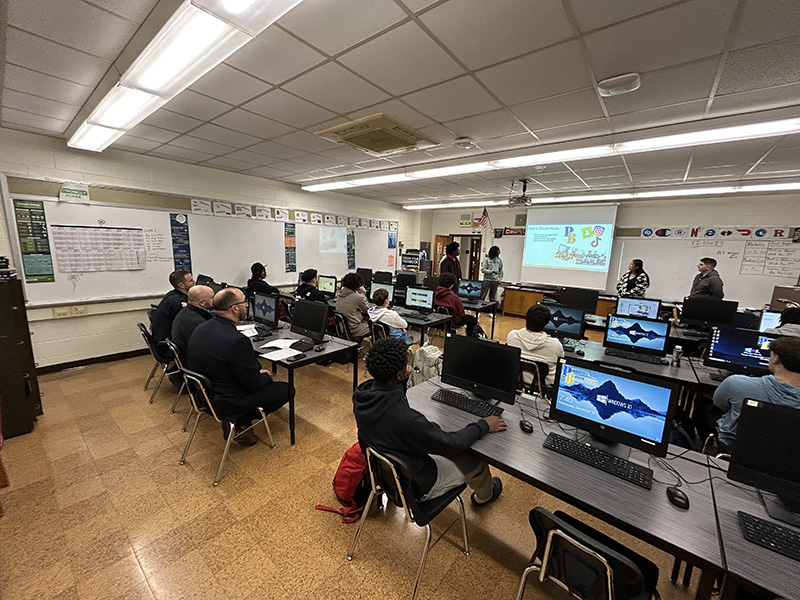 From the back of a classroom, high school students make a presentation using a screen at the front of the room to their peers and three police officers sitting in the back.