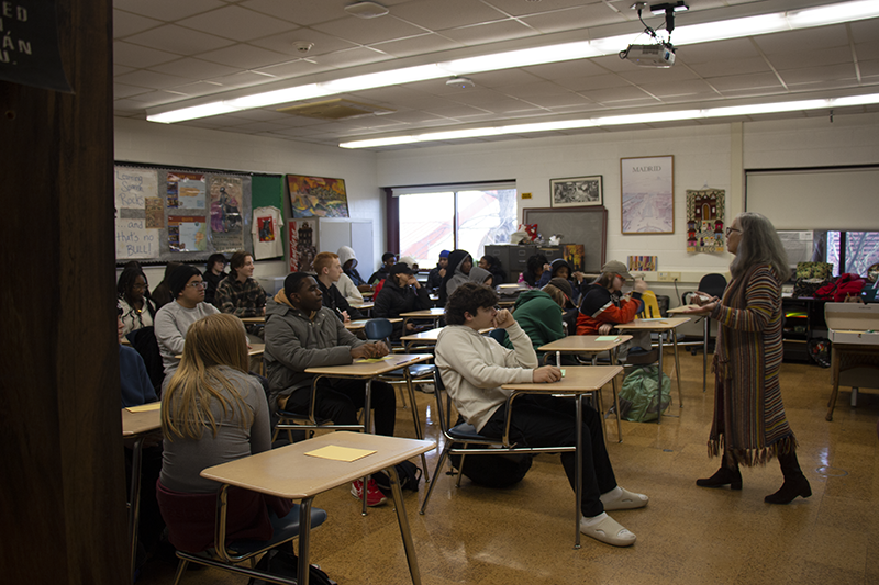 A woman in a long sweater talks to a full classroom of high school kids.